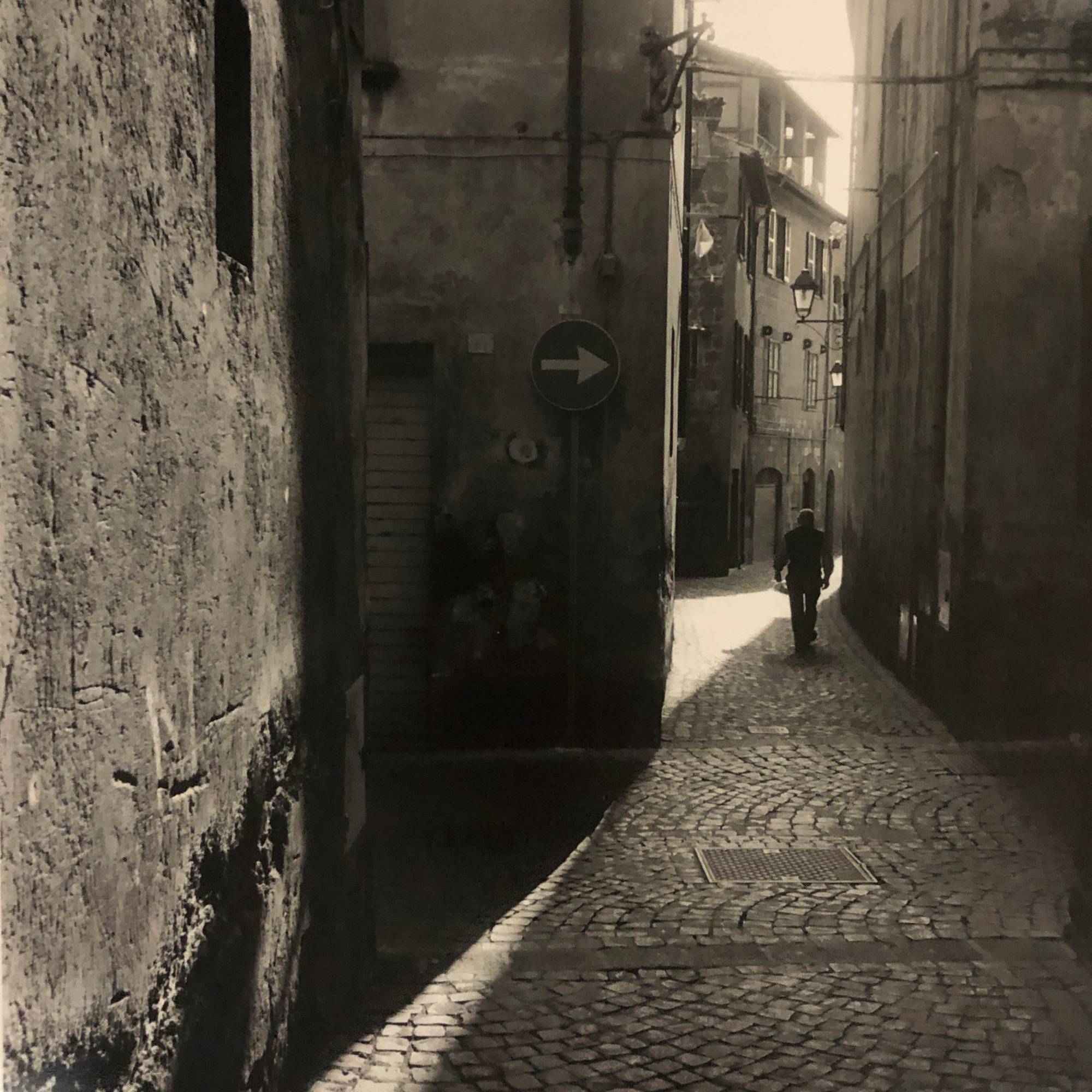 black and white photograph of man's silhouette walking away from viewer in cobblestone alley